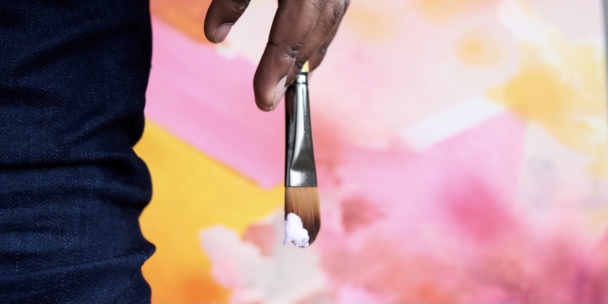 A close-up shot of a Black man’s hand, holding a paintbrush dipped in white paint, against a pink pastel backdrop.