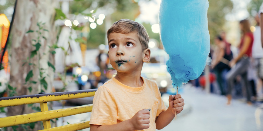 A young white toddler sits on an amusement park bench holding a giant bright blue cotton candy. He’s looking off camera with a blue stain on his mouth.
