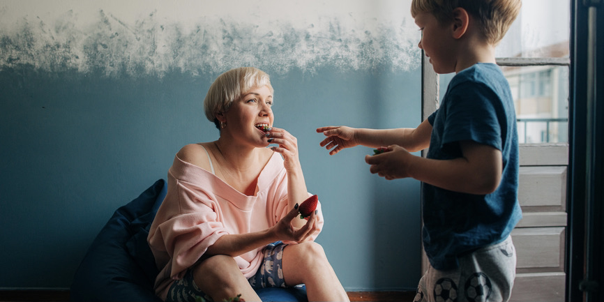 A young white mother with short blond hair wearing a pink sweatshirt sits on a beanbag against a blue wall. She’s sharing strawberries with her 5-year old son. They have short blond hair and wear a blue t-shirt.