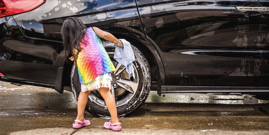A young girl with long brown hair wearing a rainbow colored dress and sandals faces away from the camera and leans the wheel of a black car.