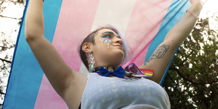 A color photograph of a trans person wearing a white tank top over a colorful body suit holds a Trans Pride flag over their head, smiling gently as they look away from the camera.