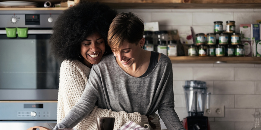 Two women—one Black with a black Afro, wearing a tan sweater, the other a white with short red hair wearing a gray long-sleeved shirt—embrace in a kitchen, smiling.