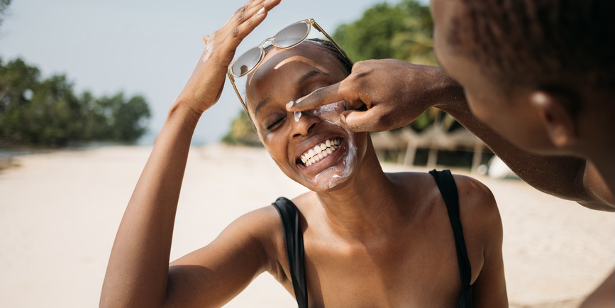 A young Black woman with short hair wearing a black bathing suit and sunglasses on her head laughs as a young Black man almost off camera dots her nose with sunscreen.