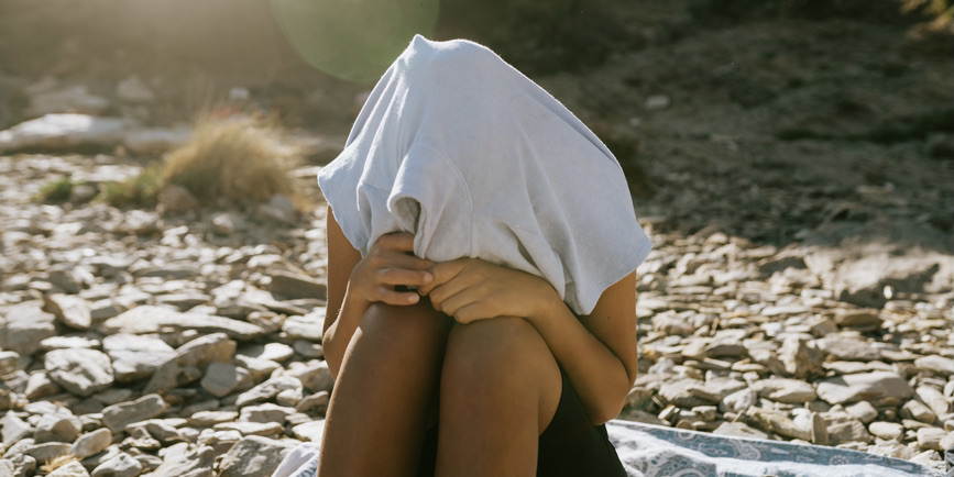 A young person with brown skin sitting on a beach blanket on a rocky beach has a t-shirt entirely covering their head.