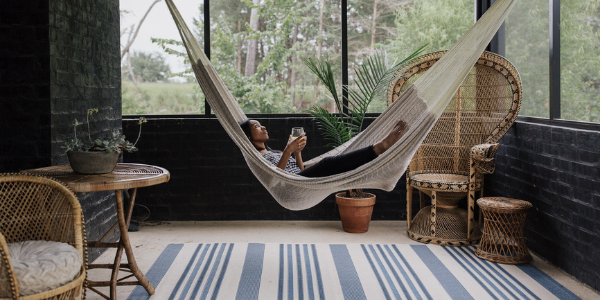 A color photo of a woman with brown skin swinging lazily in a striped hammock on a sunlit back porch.