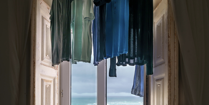 A shadowy photo of laundry hanging in a white-shuttered window with a blue horizon beyond.
