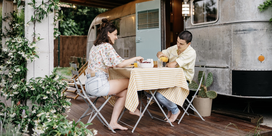 A young white couple sits outside a steel camber van, having a meal on a cloth-covered folding table.