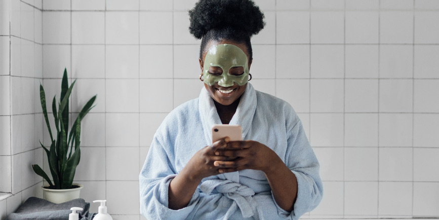 A young Black woman wearing a robe and a skincare face mask sits in the bathroom on the edge of the tub, using and smiling down at her cell phone.