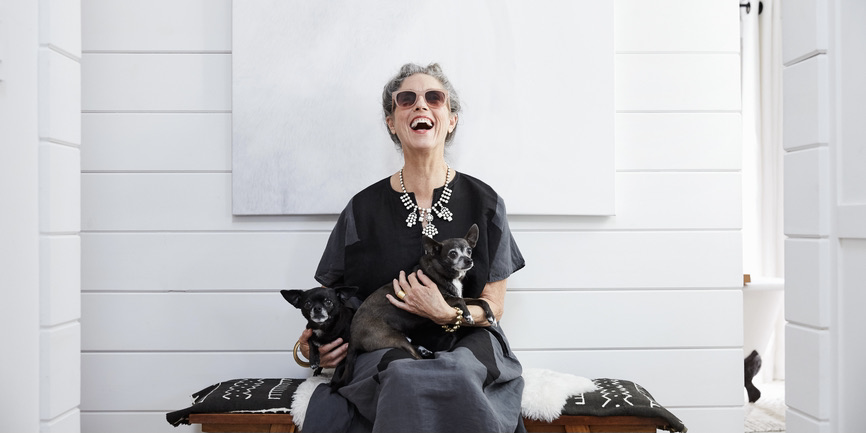 Portrait of stylish white senior woman wearing black and white clothing and sunglasses, laughing while she holds her pet dogs while seated on a bench at home.