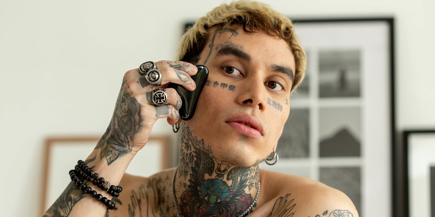 Portrait of a heavily tattooed man with light brown skin and bleached blond hair using a gua sha stone on their face.