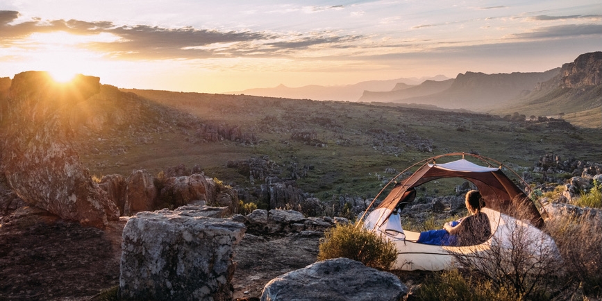 A wide view of a stunning mountain landscape and valley at sunset, and a woman with her back to the camera staring at the sunset from an open tent.