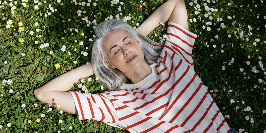 Top view of peaceful middle aged female with gray hair holding hands behind head and closing eyes while resting on blooming grass