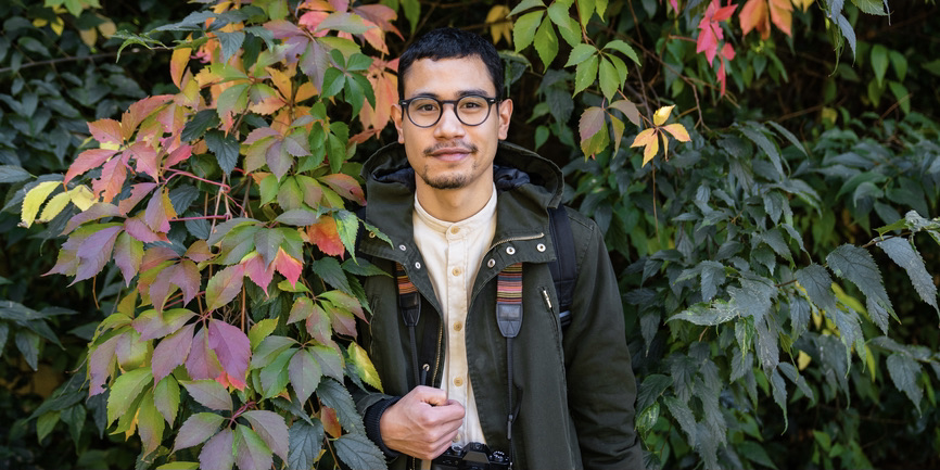 Portrait of a Filipino young man with round glasses wearing a green parka, looking at camera near some autumnal colored plants.