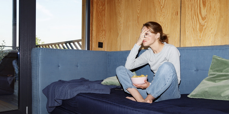 Full body photo of young white woman in a casual outfit sitting on blue sofa with crossed legs hiding face behind hand while watching scary movie and eating chips from bowl
