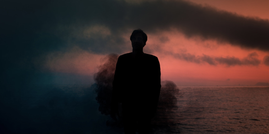 Surreal portrait of an anonymous burning man inside a dark smoke cloud during sunset near the ocean