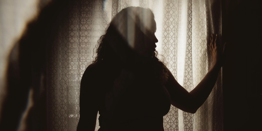 A beautiful plus size woman in her twenties stands in shadow in front of a sepia toned gauzy window curtain. 