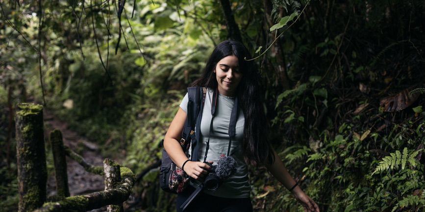 A young Latina woman with long black hair wearing a gray t-shirt and camera slung around her neck looks down and smiles as she walks through a lush tropical woodland.
